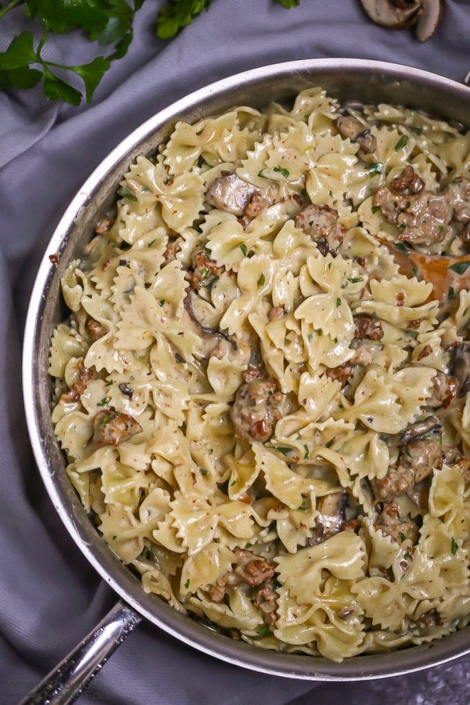 Close up of creamy farfalle pasta tossed with mushrooms and sausage in a creamy white wine sauce, in a large saute pan with a wooden spoon.