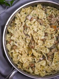 Close up of creamy farfalle pasta tossed with mushrooms and sausage in a creamy white wine sauce, in a large saute pan with a wooden spoon.