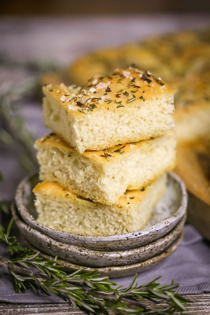 Three squares of Fluffy Focaccia Bread stacked on a little grey late with a couple sprigs of rosemary alongside.