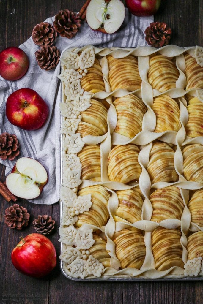 Overhead shot of Caramel Apple Slab Pie before it's baked on a half sheet tray decorated with pastry leaves and twists with apples and pinecones scattered around.