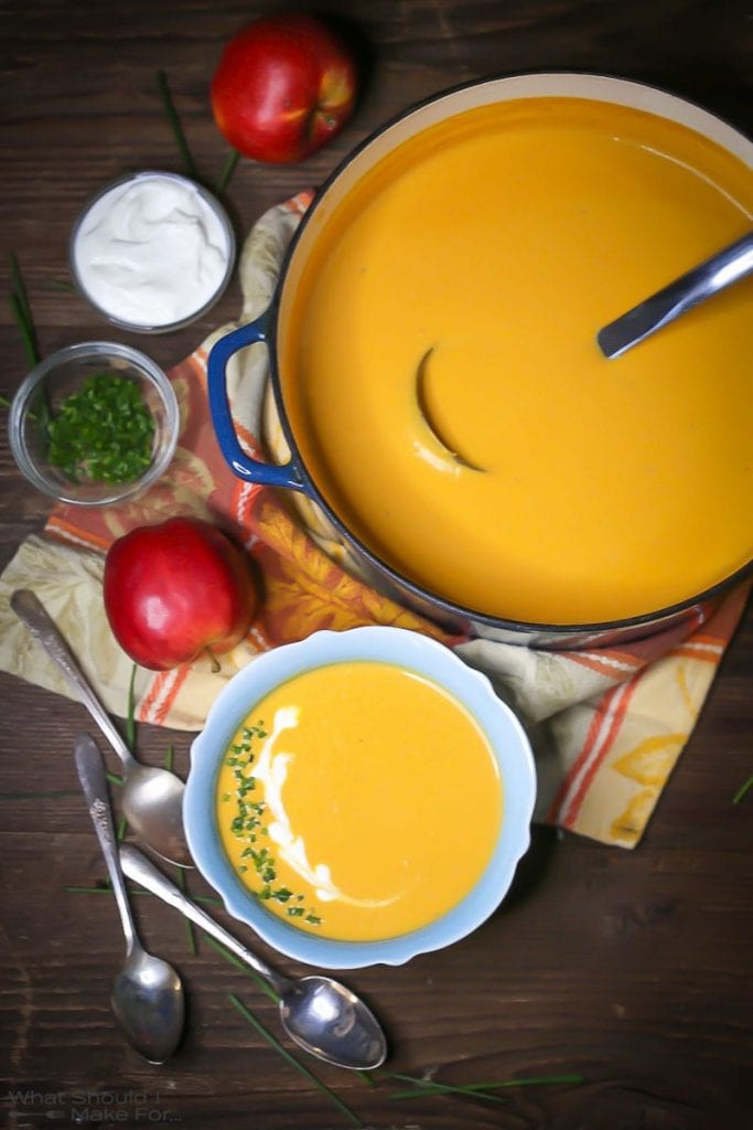 Overhead shot of Roasted Butternut Squash and Apple Soup in a blue bowl garnished with creme fraiche and chives, with spoons, an apple, and garnishes scattered around.