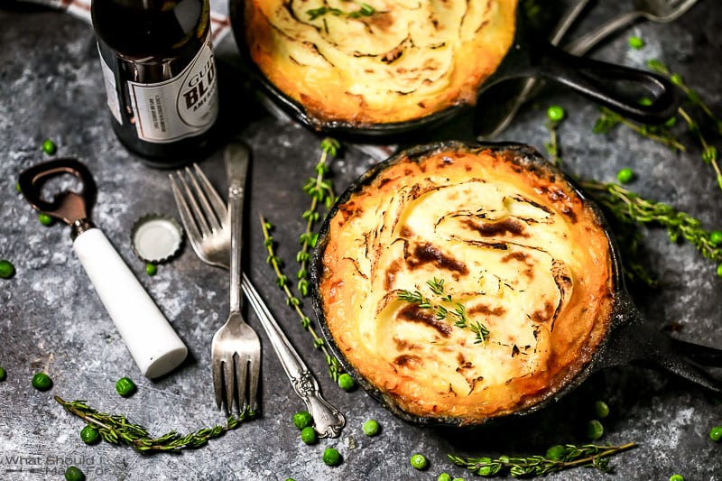 A shot of two individual servings of Shepherd's Pie in cast iron skillets with a golden crispy edged potato topping with thyme, peas, forks, and an open bottle of beer scattered around.