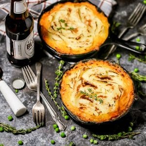 A shot of two individual servings of Shepherd's Pie in cast iron skillets with a golden crispy edged potato topping with thyme, peas, forks, and an open bottle of beer scattered around.