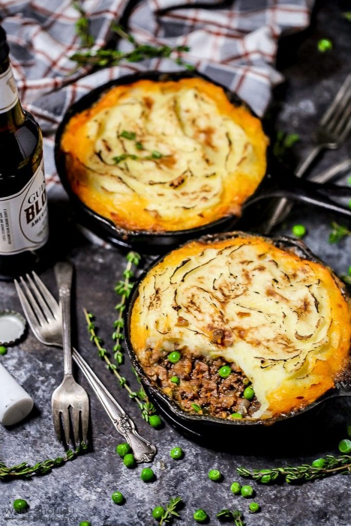 A shot of two individual servings of Shepherd's Pie in cast iron skillets with a few bites eaten, a golden crispy edged potato topping with thyme, peas, forks, and an open bottle of beer scattered around.