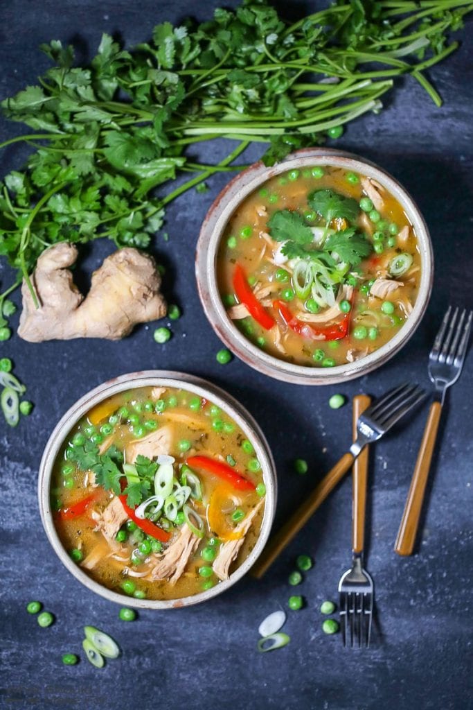 Two servings of Thai-Style Coconut Chicken Curry garnished with scallions, in a round bowl on a plate, with peas, ginger root, cilantro, and forks scattered on the table.