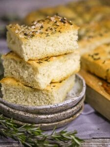Three squares of Fluffy Focaccia Bread stacked on a little grey late with a couple sprigs of rosemary alongside.