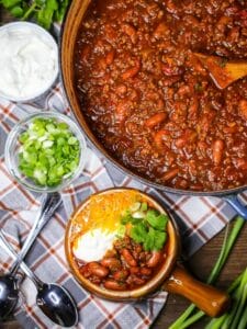 Overhead shot of Chili con Carne in a pot with a serving of chili alongside topped with sour cream, cheese, cilantro, and scallions.