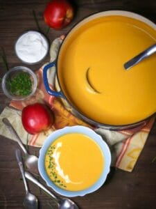 Overhead shot of Roasted Butternut Squash and Apple Soup in a blue bowl garnished with creme fraiche and chives, with spoons, an apple, and garnishes scattered around.