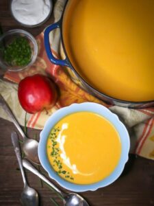 Overhead shot of Roasted Butternut Squash and Apple Soup in a blue bowl garnished with creme fraiche and chives, with spoons, an apple, and garnishes scattered around.