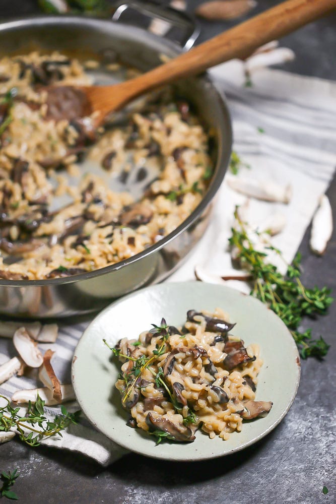 A small plate with a serving of creamy mushroom risotto in front of a silver saute pan with a wooden spoon.