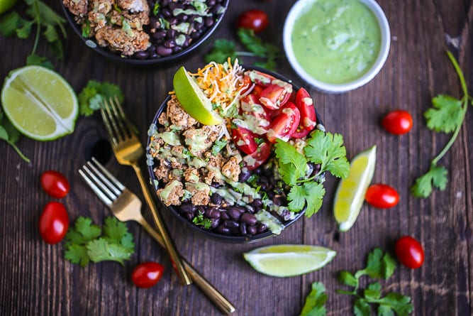 Chicken Burrito Bowls with Avocado Dressing drizzled over the top, garnished with lime and cilantro. Two gold forks are alongside the bowl and bean, avocado dressing, grape tomato, lime wedges and cilantro are scattered around.