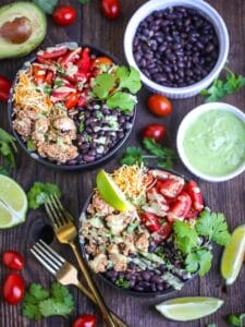 Chicken Burrito Bowls with Avocado Dressing drizzled over the top, garnished with lime and cilantro. Two gold forks are alongside the bowl and bean, avocado dressing, grape tomato, lime wedges and cilantro are scattered around.