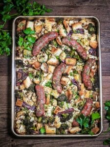 Overhead shot of Sausage Sheet Pan Dinner with Cauliflower and Broccoli on a dark wood table with fresh parsley sprigs in the corner.