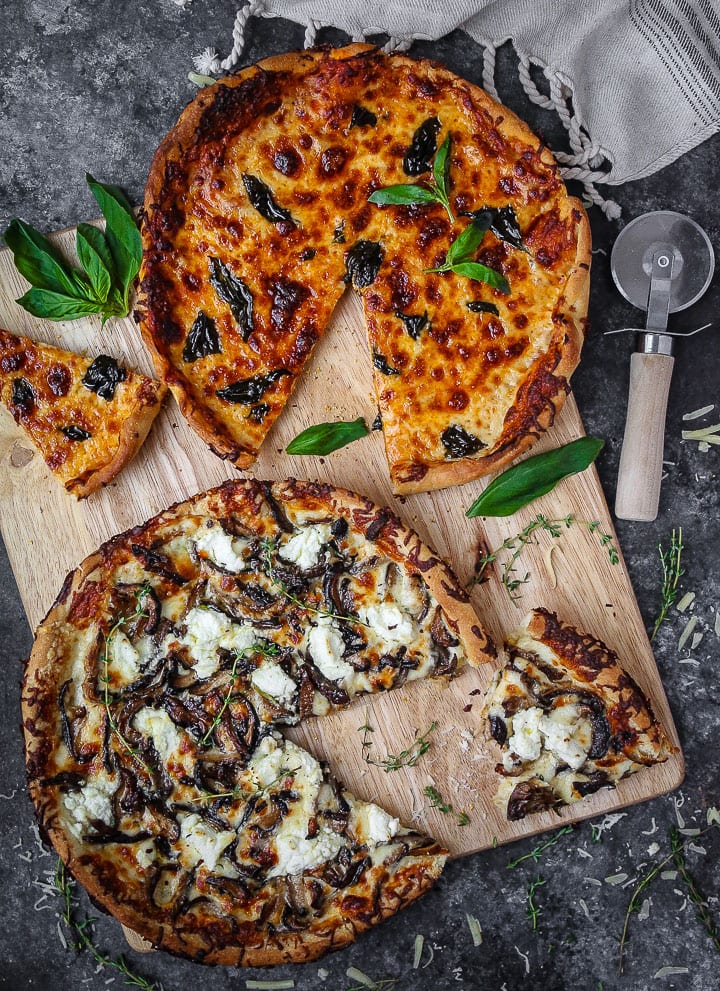 White mushroom and classic skillet pizzas on a cutting board with slices cut out, scatter with herbs and a pizza cutter laying on the table.