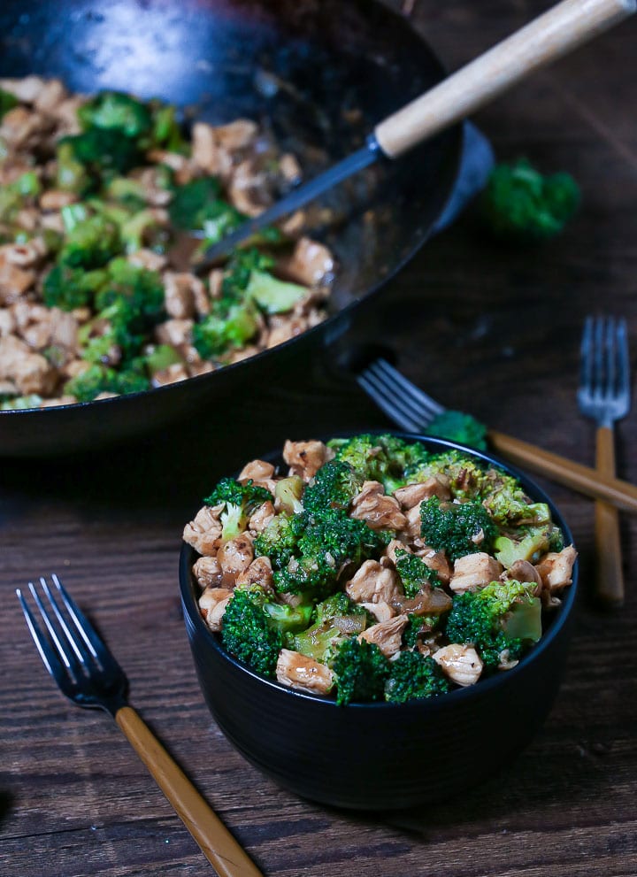 A serving of Chicken and Broccoli Stir-Fry in a black bowl with a few forks on the table and the wok in the background.