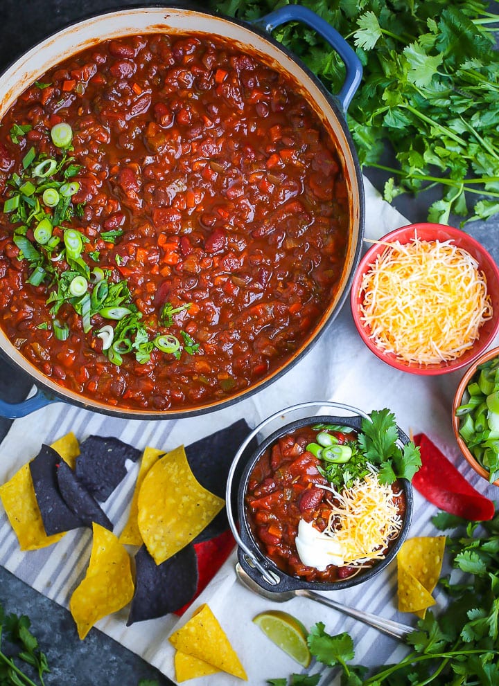 Overhead shot of Hearty Vegetarian Chili. The chili is in a large pot sprinkled with herbs and a serving of chili is in a small bowl with cheese, chips and cilantro scattered around.