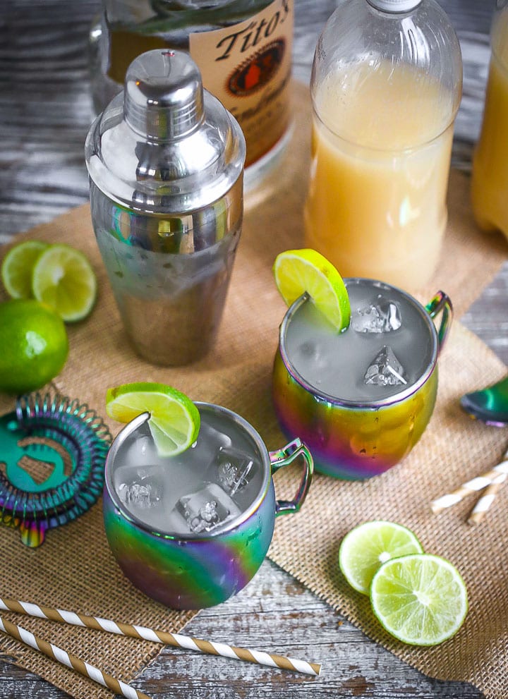 Overhead shot of Moscow Mules served in rainbow mugs with lime slices with a cocktail shaker, striped straws, ginger beer, and vodka scattered around.