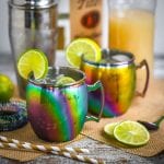 Two Moscow Mules served in rainbow mugs with lime slices with a cocktail shaker, striped straws, ginger beer, and vodka scattered around.