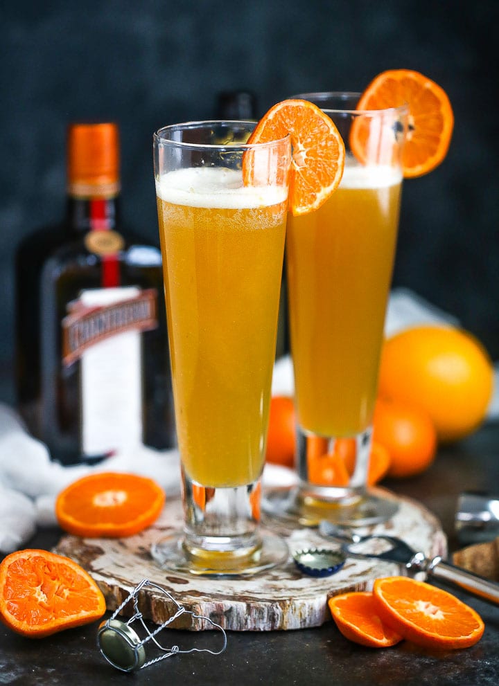 Two glasses of the The Mustache (aka the Manly Mimosa) garnished with orange slices, and orange slices, a champagne top and the bottle of cointreau are around.