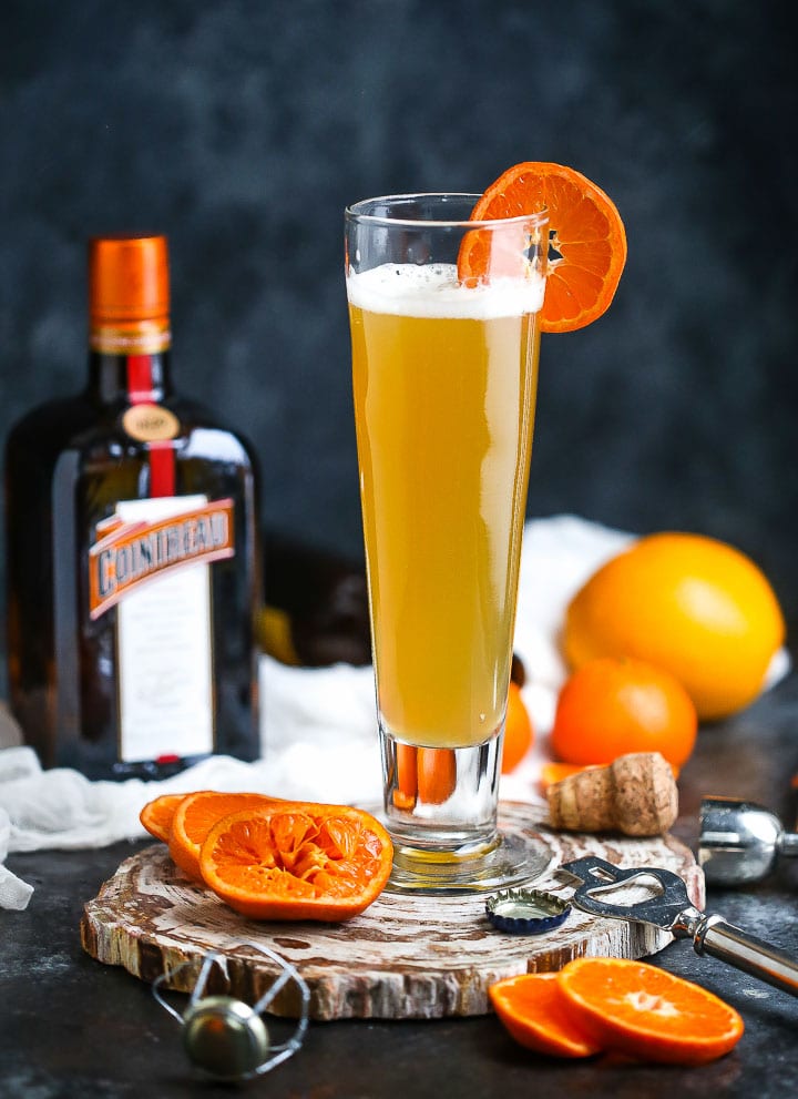 A glass of the The Mustache (aka the Manly Mimosa) garnished with orange slices, and orange slices, a champagne top and the bottle of cointreau are around.