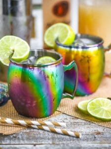 Two Moscow Mules served in rainbow mugs with lime slices with a cocktail shaker, striped straws, ginger beer, and vodka scattered around.