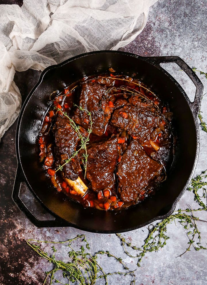 Irish Whiskey Braised Short Ribs in a cast iron dutch oven with thyme sprigs scattered around and a white linen towel draped in the corner.