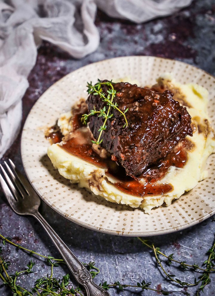 A serving of Irish Whiskey Braised Short Ribs over mashed potatoes on a polka dotted plate with thyme sprigs and a fork on the table.