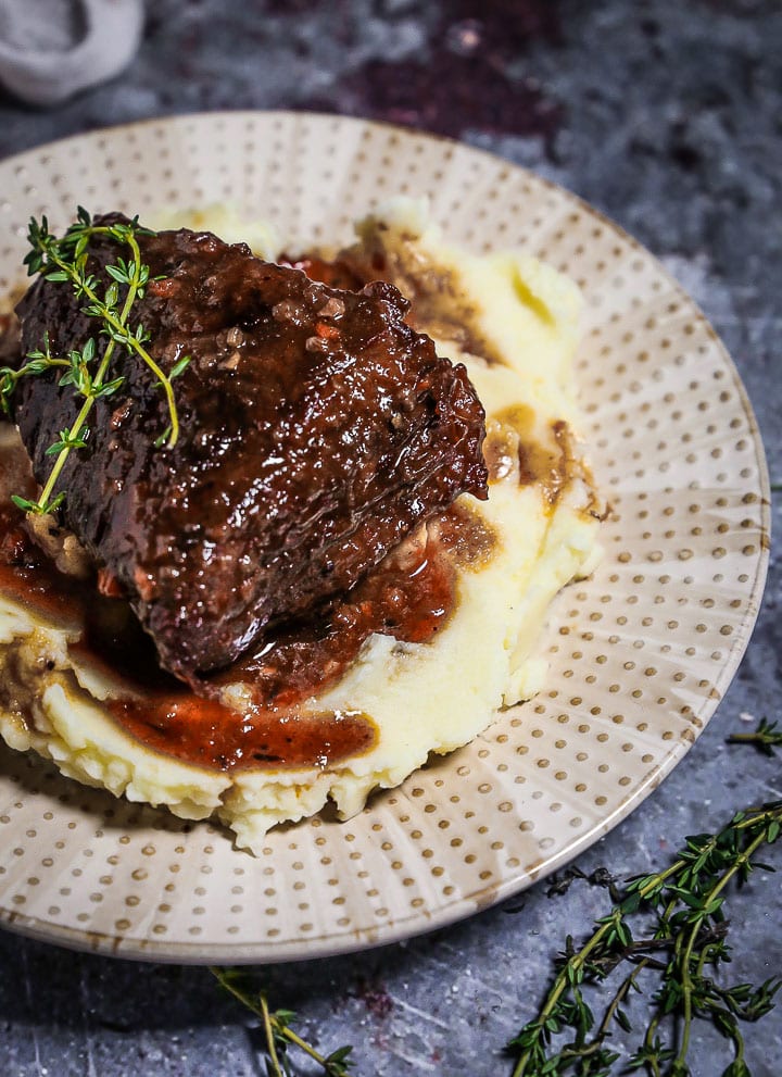 A serving of Irish Whiskey Braised Short Ribs over mashed potatoes on a polka dotted plate with thyme sprigs on the table.
