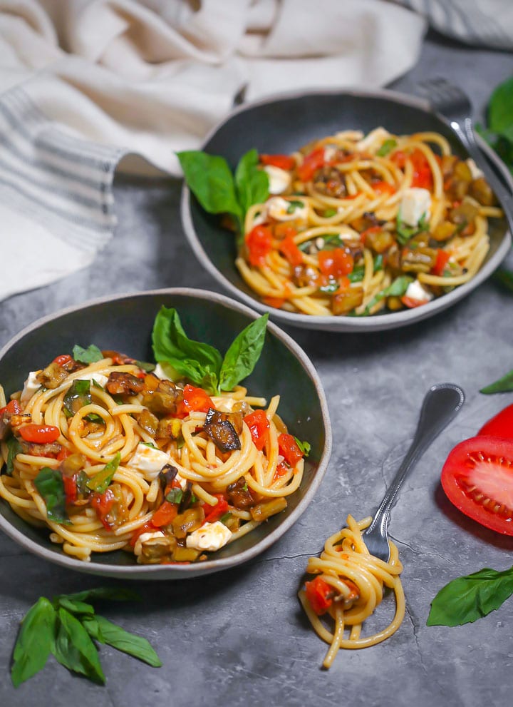 Two black bowls of roasted eggplant pasta with a few basil leaves garnishing the bowls and a fork with a twirl of pasta alongside.
