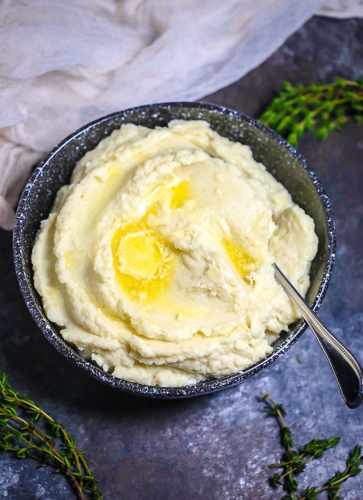 Creamy Cauliflower Mash topped with melted butter in a black speckled bowl with a spoon in it and sprigs of thyme scattered on the table.