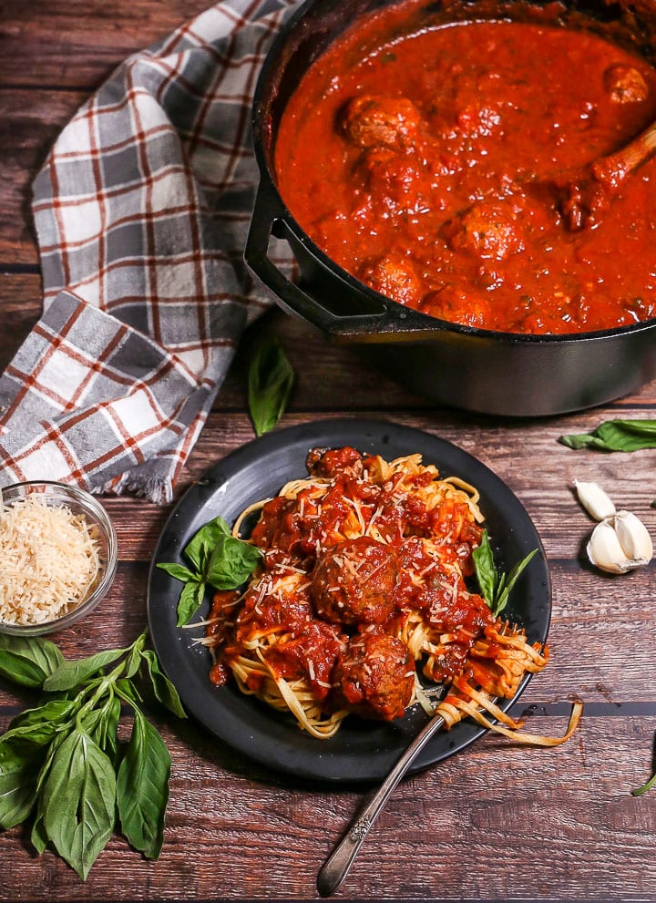 A serving of Sunday Sauce and Meatballs over fettuccine on a black plate garnished with fresh basil and a fork twirled in the pasta. The pot of sauce is in the corner.