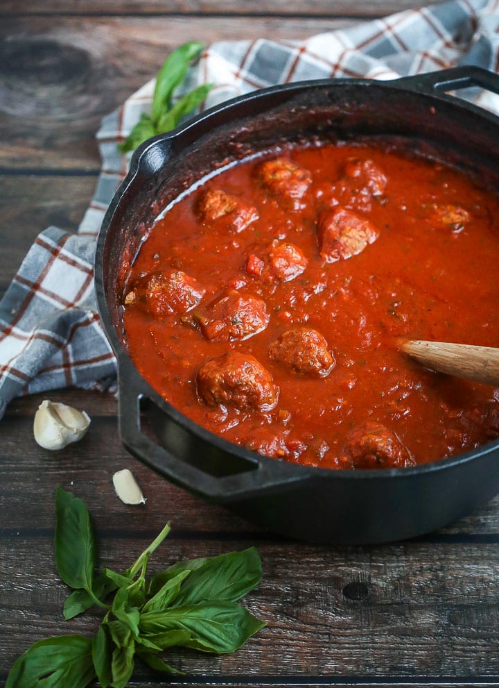 A pot of Sunday Sauce and Meatballs with a wooden spoon in the pot, a couple garden cloves and fresh basil alongside.