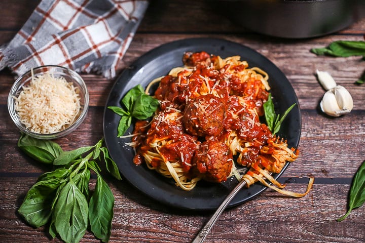 A serving of Sunday Sauce and Meatballs over fettuccine on a black plate garnished with fresh basil and a fork twirled in the pasta.