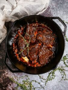 Irish Whiskey Braised Short Ribs in a cast iron dutch oven with thyme sprigs scattered around and a white linen towel draped in the corner.