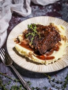 A serving of Irish Whiskey Braised Short Ribs over mashed potatoes on a polka dotted plate with thyme sprigs and a fork on the table.