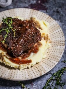 A serving of Irish Whiskey Braised Short Ribs over mashed potatoes on a polka dotted plate with thyme sprigs on the table.