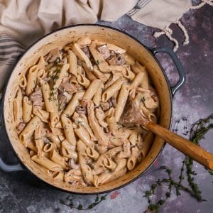 Creamy Goat Cheese Pasta with Chicken and Mushrooms in a dutch oven with a wooden spoon in to stir, thyme sprigs scattered around and beige blue striped dish towel.