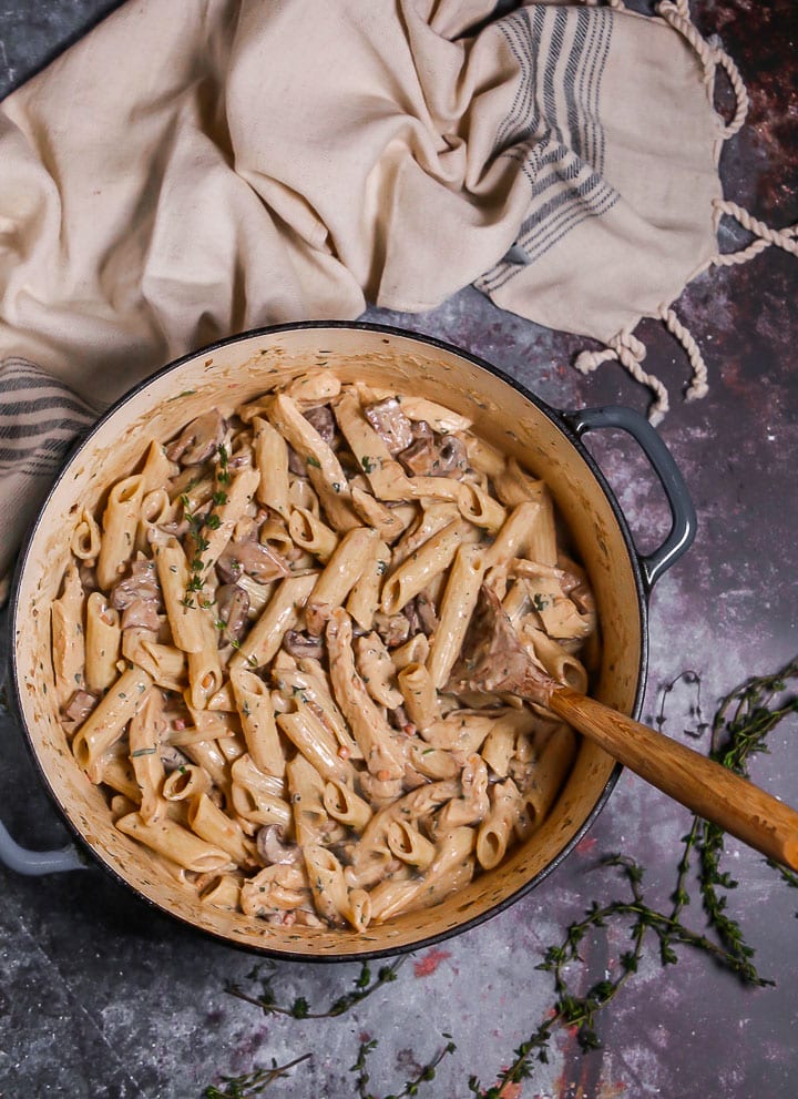 Creamy Goat Cheese Pasta with Chicken and Mushrooms in a dutch oven with a wooden spoon in to stir, thyme sprigs scattered around and beige blue striped dish towel.