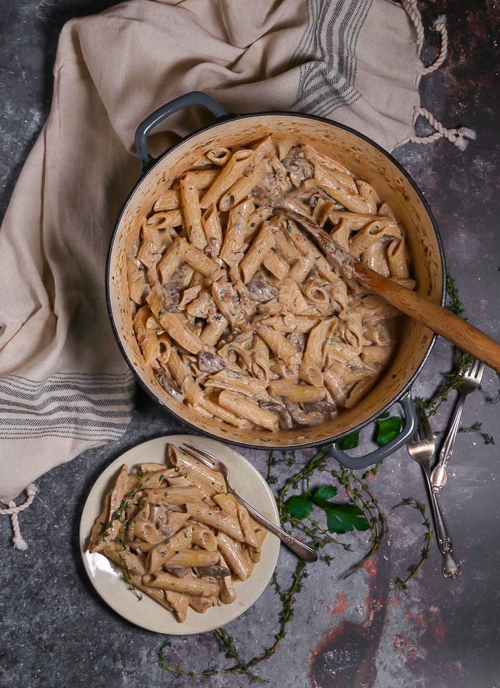 Overhead shot of Creamy Goat Cheese Pasta with Chicken and Mushrooms in a pot with a wooden spoon and a serving on a small plate with forks and thyme scattered around.