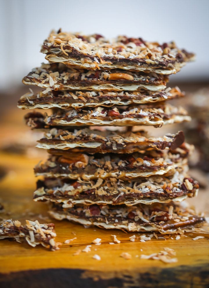 Toffee Matzo Brittle (aka matzo crack), is topped with toasted coconut and pecans is stacked on top of a wood board.