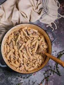 Creamy Goat Cheese Pasta with Chicken and Mushrooms in a dutch oven with a wooden spoon in to stir, thyme sprigs scattered around and beige blue striped dish towel.