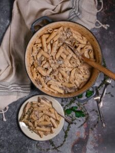 Overhead shot of Creamy Goat Cheese Pasta with Chicken and Mushrooms in a pot with a wooden spoon and a serving on a small plate with forks and thyme scattered around.