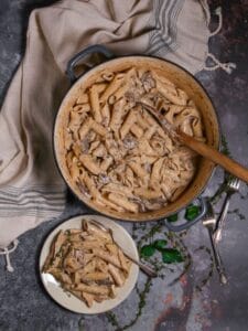 Overhead shot of Creamy Goat Cheese Pasta with Chicken and Mushrooms in a pot with a wooden spoon and a serving on a small plate with forks and thyme scattered around.