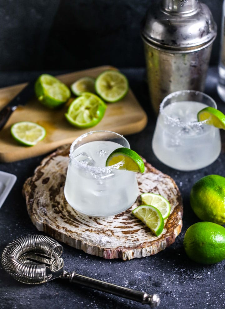 Two margaritas garnished with salt and lime slices, with a cocktail shaker, strainers and limes scattered around.