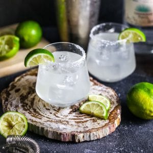 Two margaritas garnished with salt and lime slices, with a cocktail shaker, strainers and limes scattered around.