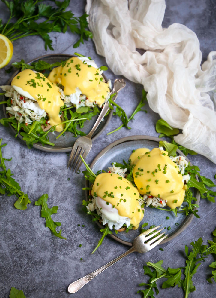 Two servings of Crab Eggs Benedict on round blue plates garnished with minced chives, with forks, fresh arugula and a white gauzy towel on the table.
