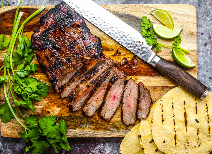 Grilled and rested carne asada, being sliced on a cutting board with cilantro, tortillas and limes scattered around.