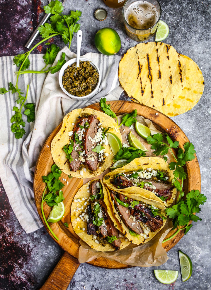 Carne Asada Tacos on a round wooden board with cilantro, limes, grilled tortillas and onion relish scattered around.