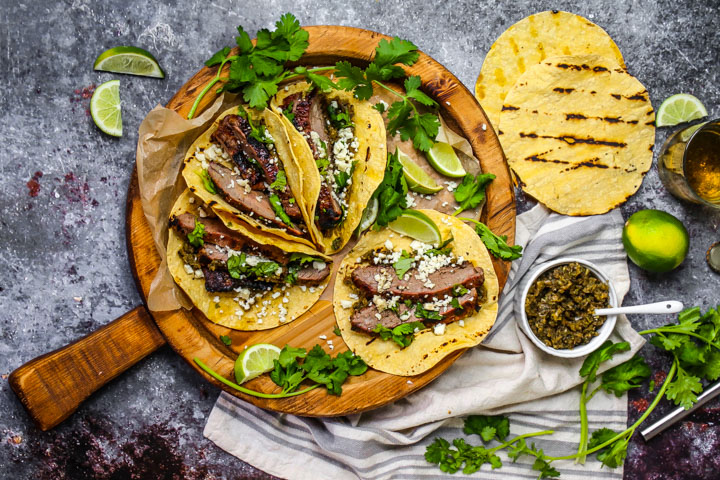 Carne Asada Tacos on a round wooden board with cilantro, limes, grilled tortillas and onion relish scattered around.