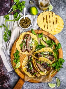 Carne Asada Tacos on a round wooden board with cilantro, limes, grilled tortillas and onion relish scattered around.